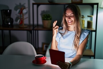 Canvas Print - Young hispanic woman using touchpad sitting on the table at night gesturing finger crossed smiling with hope and eyes closed. luck and superstitious concept.