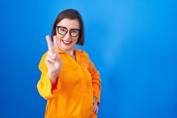 Poster - Middle age hispanic woman wearing glasses standing over blue background smiling looking to the camera showing fingers doing victory sign. number two.