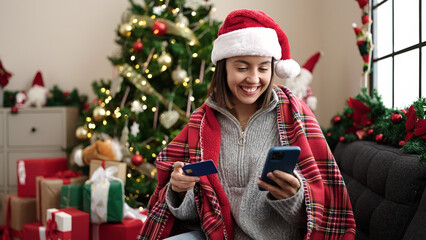 Poster - Young beautiful hispanic woman using smartphone and credit card sitting on sofa by christmas tree at home