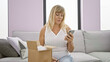 © Krakenimages.com - Beautiful middle-aged blonde woman seriously unpacking a box at home, texting on her smartphone while relaxed on the living room sofa