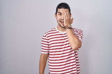 Wall Mural - Hispanic man with long hair standing over isolated background covering one eye with hand, confident smile on face and surprise emotion.