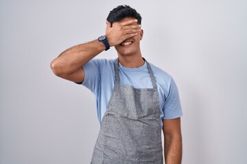 Sticker - Hispanic young man wearing apron over white background smiling and laughing with hand on face covering eyes for surprise. blind concept.