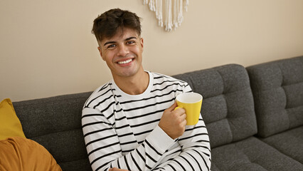 Poster - Captivating glimpse into coffee-filled mornings, as a handsome young hispanic man, radiating positivity and confidence, luxuriates on a comfy living room sofa, savoring every sip in his cozy apartment