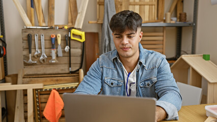 Poster - Serious-faced young hispanic man, a skilled carpenter, engrossed in his laptop amidst a busy carpentry workshop, presenting the blend of traditional woodwork and modern technology.