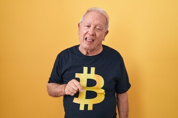 Poster - Senior man with grey hair wearing bitcoin t shirt sticking tongue out happy with funny expression. emotion concept.