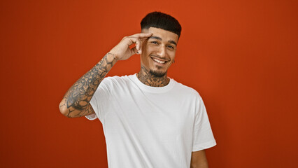 Handsome, tattooed young latin man confidently giving a military salutation, a joyful smile playing on his face. frozen in a casual pose over a fiery red isolated background.