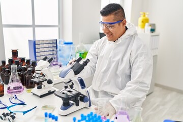 Wall Mural - Young latin man scientist wearing covid protection uniform using microscope at laboratory