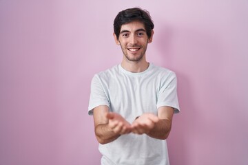 Poster - Young hispanic man standing over pink background smiling with hands palms together receiving or giving gesture. hold and protection