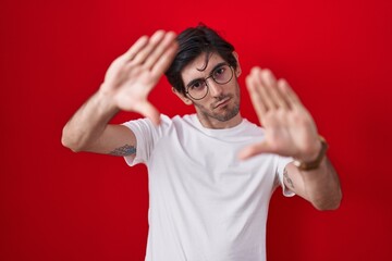 Poster - Young hispanic man standing over red background doing frame using hands palms and fingers, camera perspective