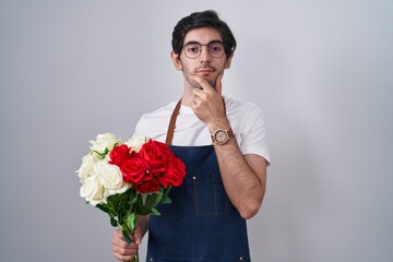 Wall Mural - Young hispanic man holding bouquet of white and red roses with hand on chin thinking about question, pensive expression. smiling with thoughtful face. doubt concept.