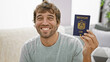 Chilled-out young man, smiling happily with confidence, casually sitting on sofa at home, holding his travel passport of united arab emirates, ready to enjoy his fun vacation trip
