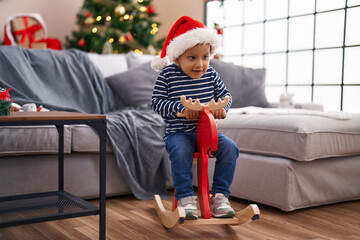 Sticker - Adorable hispanic toddler playing with reindeer rocking by christmas tree at home