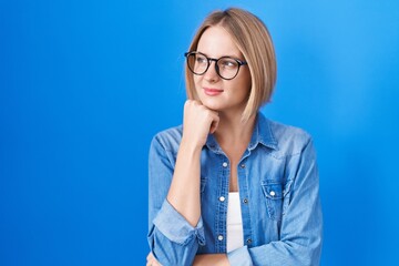 Wall Mural - Young caucasian woman standing over blue background with hand on chin thinking about question, pensive expression. smiling with thoughtful face. doubt concept.