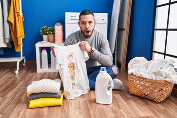 Sticker - Hispanic man holding dirty t shirt with stain afraid and shocked with surprise and amazed expression, fear and excited face.