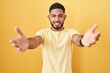 © Krakenimages.com - Young hispanic man standing over yellow background looking at the camera smiling with open arms for hug. cheerful expression embracing happiness.