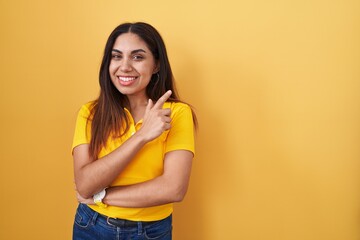 Wall Mural - Young arab woman standing over yellow background with a big smile on face, pointing with hand finger to the side looking at the camera.