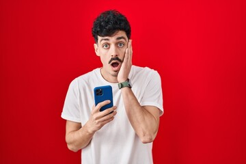 Wall Mural - Hispanic man using smartphone over red background afraid and shocked, surprise and amazed expression with hands on face