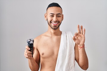 Wall Mural - Young hispanic man holding electric razor doing ok sign with fingers, smiling friendly gesturing excellent symbol