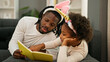 © Krakenimages.com - African american father and daughter wearing funny diadem lying on sofa reading book at home
