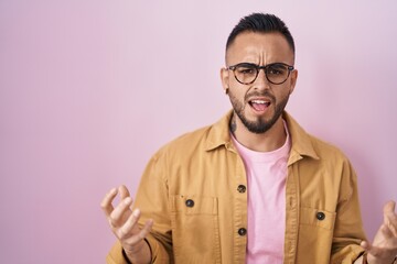 Poster - Young hispanic man standing over pink background crazy and mad shouting and yelling with aggressive expression and arms raised. frustration concept.