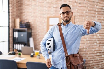 Poster - Young hispanic man working at the office holding bike helmet with angry face, negative sign showing dislike with thumbs down, rejection concept