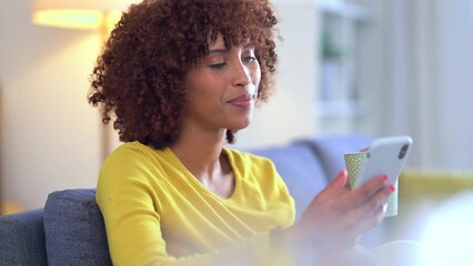 Wall Mural - Trendy young woman laughing and texting on a phone, browsing social media or surfing the internet. Female drinking a cup of coffee and banking on an app for convenience on a comfortable sofa at home