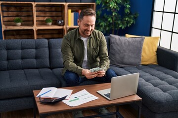 Poster - Young caucasian man using laptop counting dollars at home