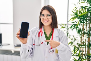 Poster - Young hispanic doctor woman showing smartphone screen pointing finger to one self smiling happy and proud