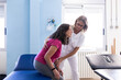 A middle aged blonde physiotherapist with glasses is helping her patient to sit down.