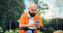 Positive Man With Bicycle Wearing Orange Shirt Texting On Phone And Looking Around Near Building Outdoors. Attractive Happy Male Tapping And Scrolling On Mobile Phone.