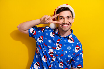 Sticker - Photo of optimistic young guy showing v sign cover face peace symbol wear blue cool santa claus shirt isolated on yellow color background