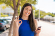 © luismolinero - Young pretty Brazilian woman using mobile phone at outdoors showing ok sign with fingers
