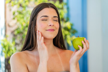 Wall Mural - Young pretty Brazilian woman at outdoors holding an apple with happy expression