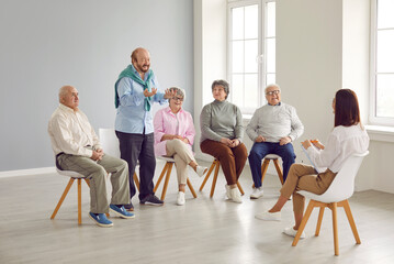 Senior people in group therapy. Happy retired old men and women sitting on chairs in a light office or retirement house and talking to a young friendly woman therapist with a clipboard