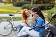 © pucko_ns - Two girl friends sitting on bench, looking at mobile phone and smile in public par with bicycle on background
