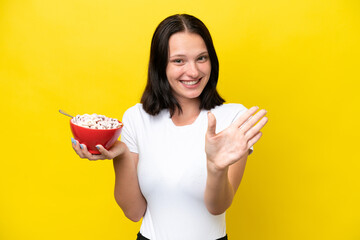 Young caucasian woman holding a bowl of cereals isolated on yellow background saluting with hand with happy expression