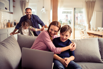 Wall Mural - Family having fun at home together. Mother and son sitting on couch using laptop while father plays with daughter in background