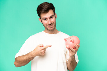 Wall Mural - Young caucasian man holding piggybank isolated on green background and pointing it