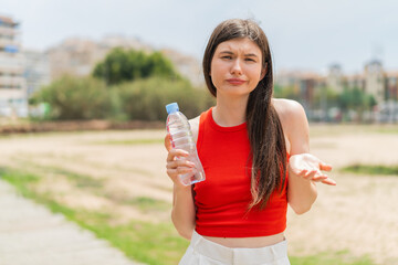 Wall Mural - Young pretty Ukrainian woman with a bottle of water at outdoors making doubts gesture while lifting the shoulders