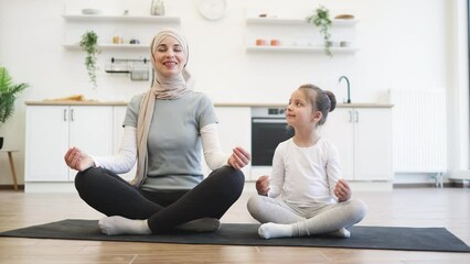 Wall Mural - Full length view of relaxed muslim female sitting in yoga pose with little child on rubber mat in modern apartment. Fit mother exercising with fingers in gyan mudra with daughter looking at each other