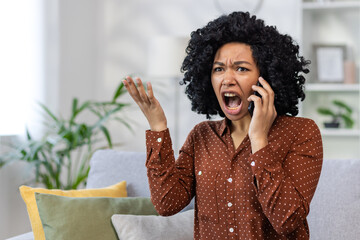 Frustrated and angry woman talking on the phone, African American woman shouting and angry at the interlocutor, sitting on the sofa in the living room of the house alone