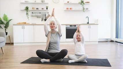 Wall Mural - Muslim female with little daughter in sports outfit and closed eyes holding folded palms overhead while meditating in crossed-legged position in fitness studio. Healthy family practicing yoga together