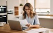 © piai - Smiling businesswoman reading financial document and using laptop on desk while working from home