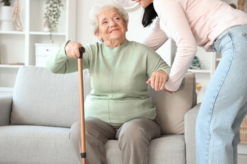 Young woman helping her mother with stick to get up from sofa at home