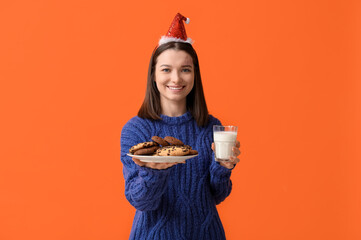 Sticker - Beautiful young woman in Christmas hat with tasty cookies and glass of milk on orange background