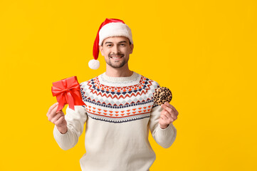 Canvas Print - Handsome man in Santa hat with gift box and tasty cookie on yellow background