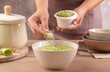 Woman adding fresh microgreens into bowl with tasty leek soup at wooden table, closeup