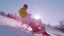 SLOW MOTION, LENS FLARE: Active Young Guy Enjoys A Sleigh Ride On Winter Holidays In The Alps. Sun Is Shining And Flying Snow Is Sparkling When He Passes By Riding On A Wooden Sled Down The Snowy Hill