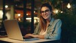 Cheerful Caucasian woman with glasses using laptop in a cozy evening café, ideal for remote work and leisure.