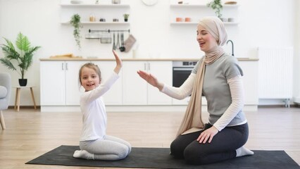 Wall Mural - Family, young muslim mother and little daughter engaged in fitness, yoga, exercise at home. Smiling woman in hijab looking at small kid girl sitting on the carpet floor on knees and giving high five.